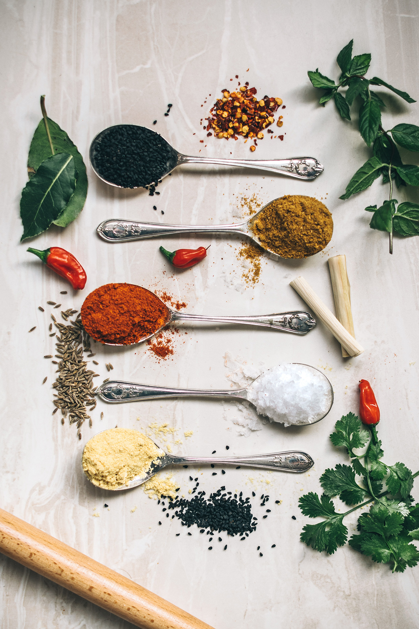 A set of five spoons on a counter with different spices in each, surrounded by small piles of other spices.